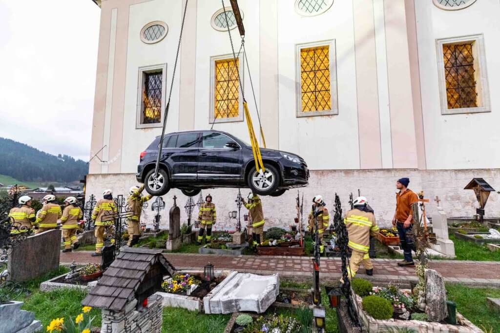 Feuerwehrleute heben ein Auto über einem Friedhof mit Grabsteinen und Blumen.
